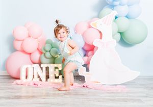 Toddler girl celebrates first birthday with pastel balloons, ONE sign, and white dress cutout backdrop.