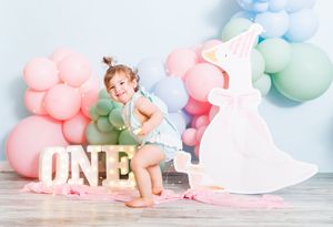 Toddler girl celebrating first birthday with colorful balloons, ONE sign, and unicorn cutout in pastel party setting.