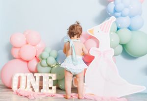 Toddler girl in blue romper at a pastel balloon first birthday party with ONE sign and swan cutout.