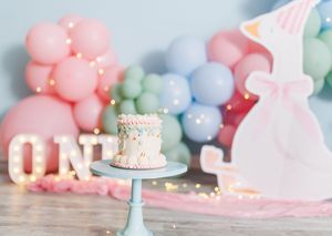 Pastel balloon garland backdrop with a small smash cake on a blue stand and marquee ONE letters for a first birthday.