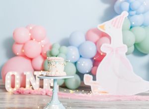 Pastel first birthday party setup with balloon garland, smash cake on stand, and unicorn cutout decoration.