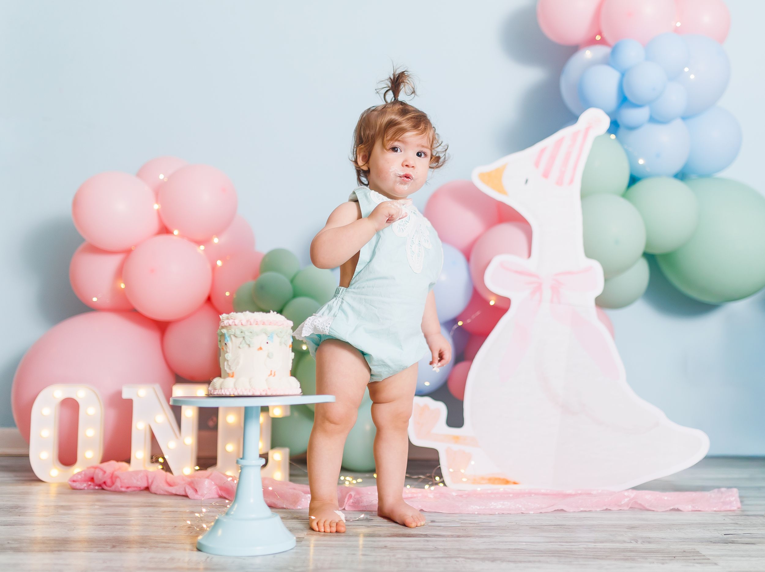 Toddler girl celebrates first birthday with pastel balloons, smash cake, and decorative number one sign.