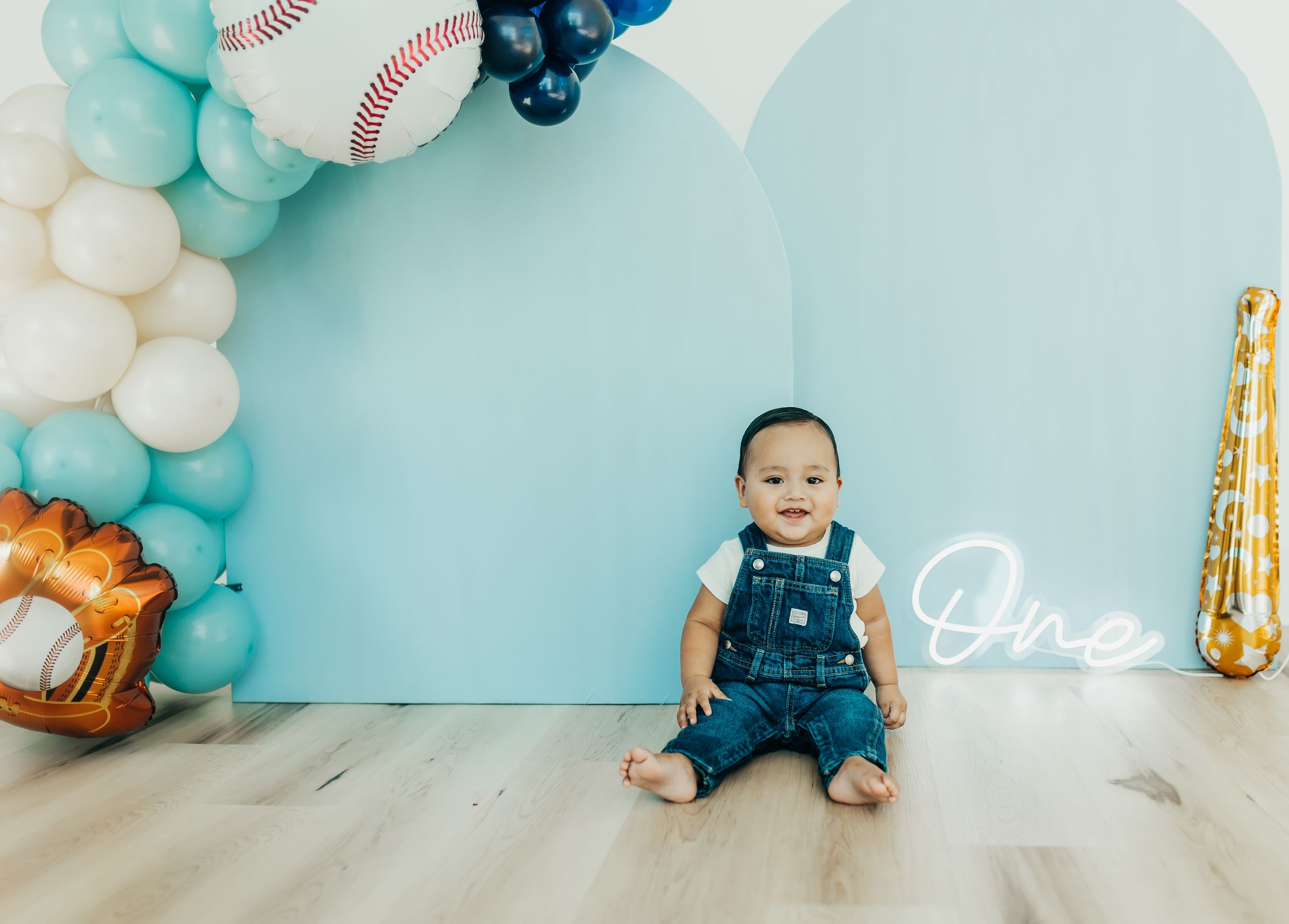 First birthday party setup with blue balloon arch, baseball decor, and neon 'One' sign against light blue backdrop.