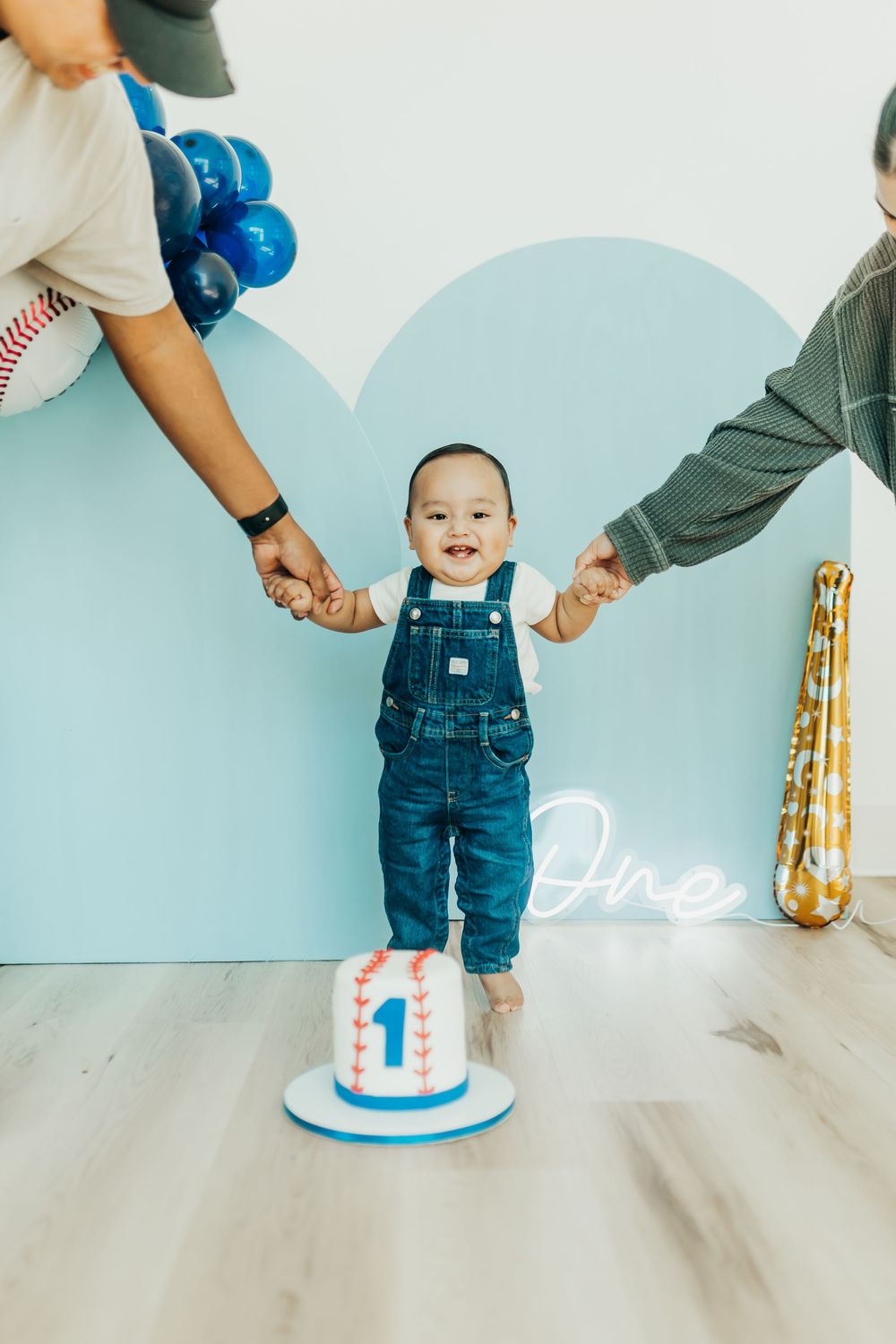 A small child in denim overalls stands between two adults while celebrating their first birthday with a cake.