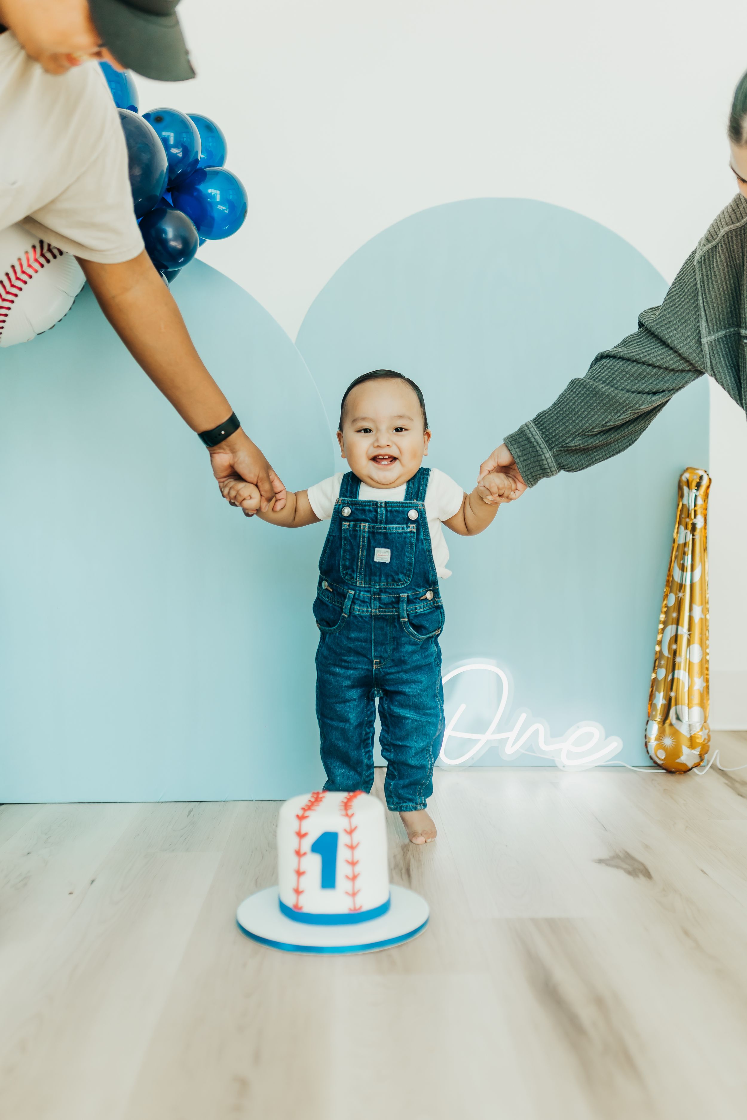 A small child in denim overalls stands between two adults while celebrating their first birthday with a cake.