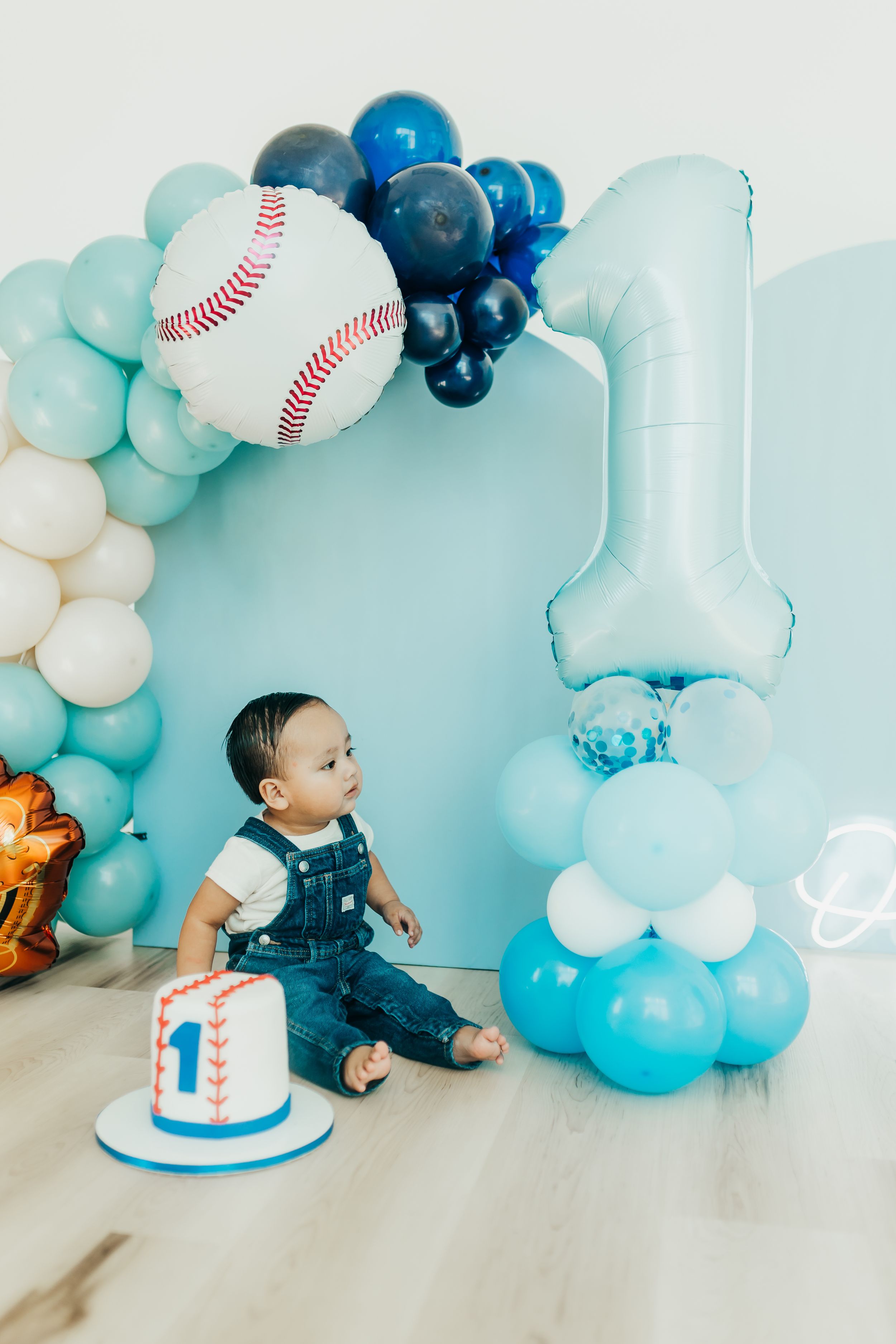 First birthday party setup with baseball theme featuring blue balloon arch, smash cake and large number one balloon.