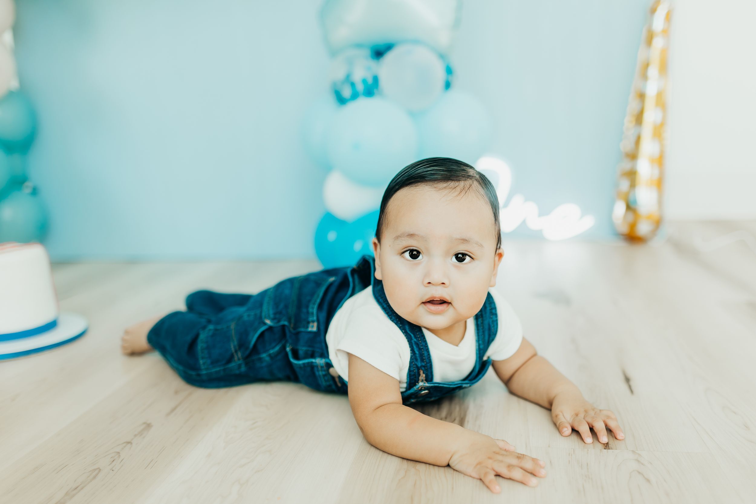A baby in a white outfit lies on a light gray backdrop with blue and gold balloons for a birthday photoshoot.