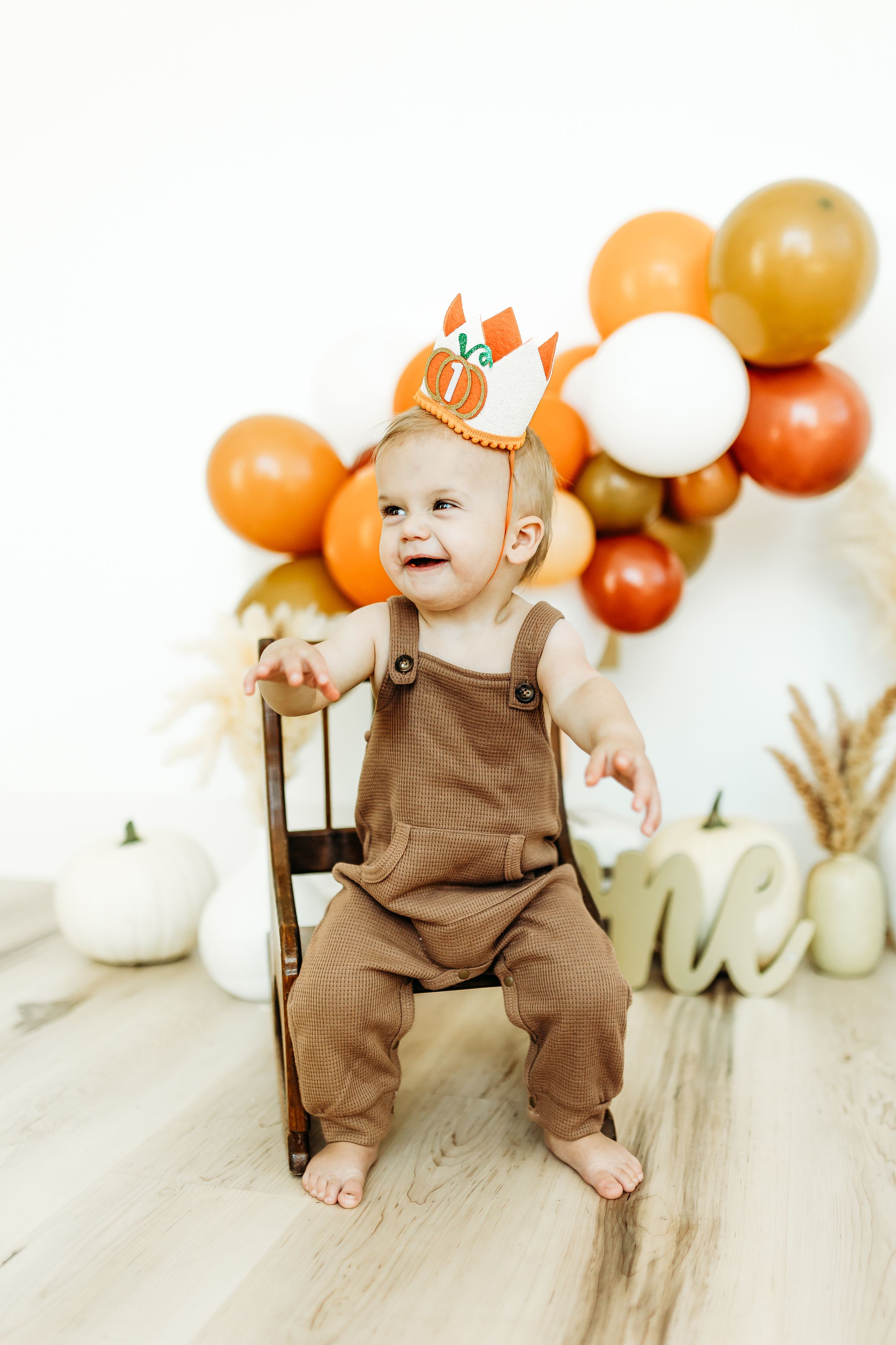 Smiling baby in brown overalls sits on wooden floor with orange and white balloon decorations in background.