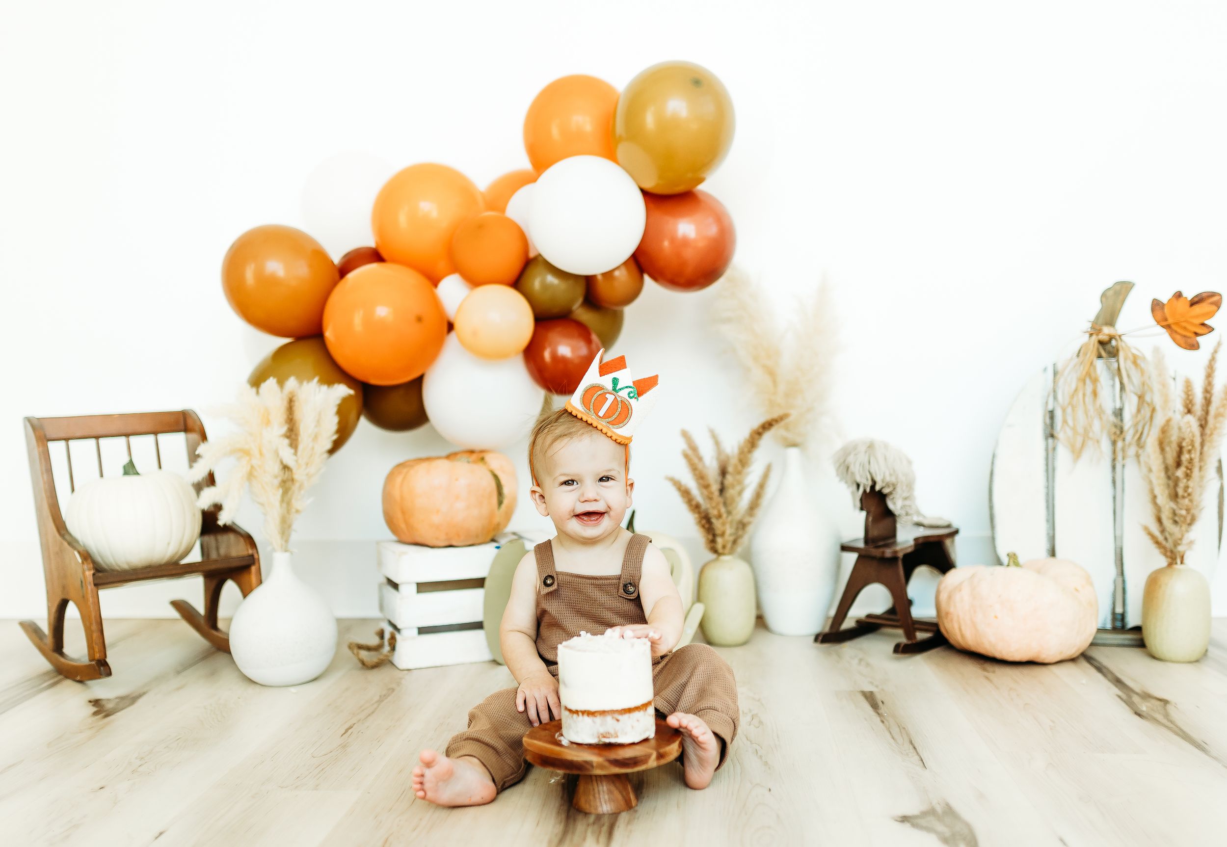 Autumnal nursery scene with orange and white balloons, pumpkins, dried flowers and wooden rocking horse on light wood floor.