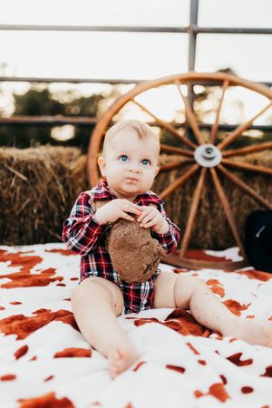 A baby in a plaid shirt sits on a white blanket with brown wagon wheels visible in the background.