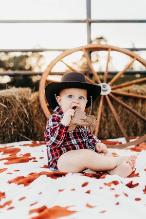 Baby in plaid shirt and black hat lies on red blanket with rustic wagon wheel backdrop.
