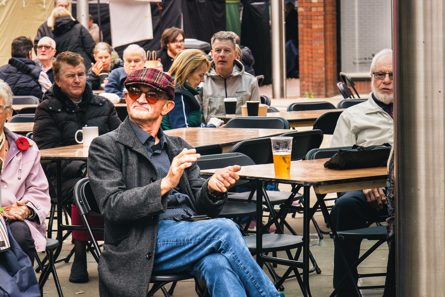 People seated at outdoor tables in urban setting, with focus on one older man wearing a flat cap that is clapping along to some music.