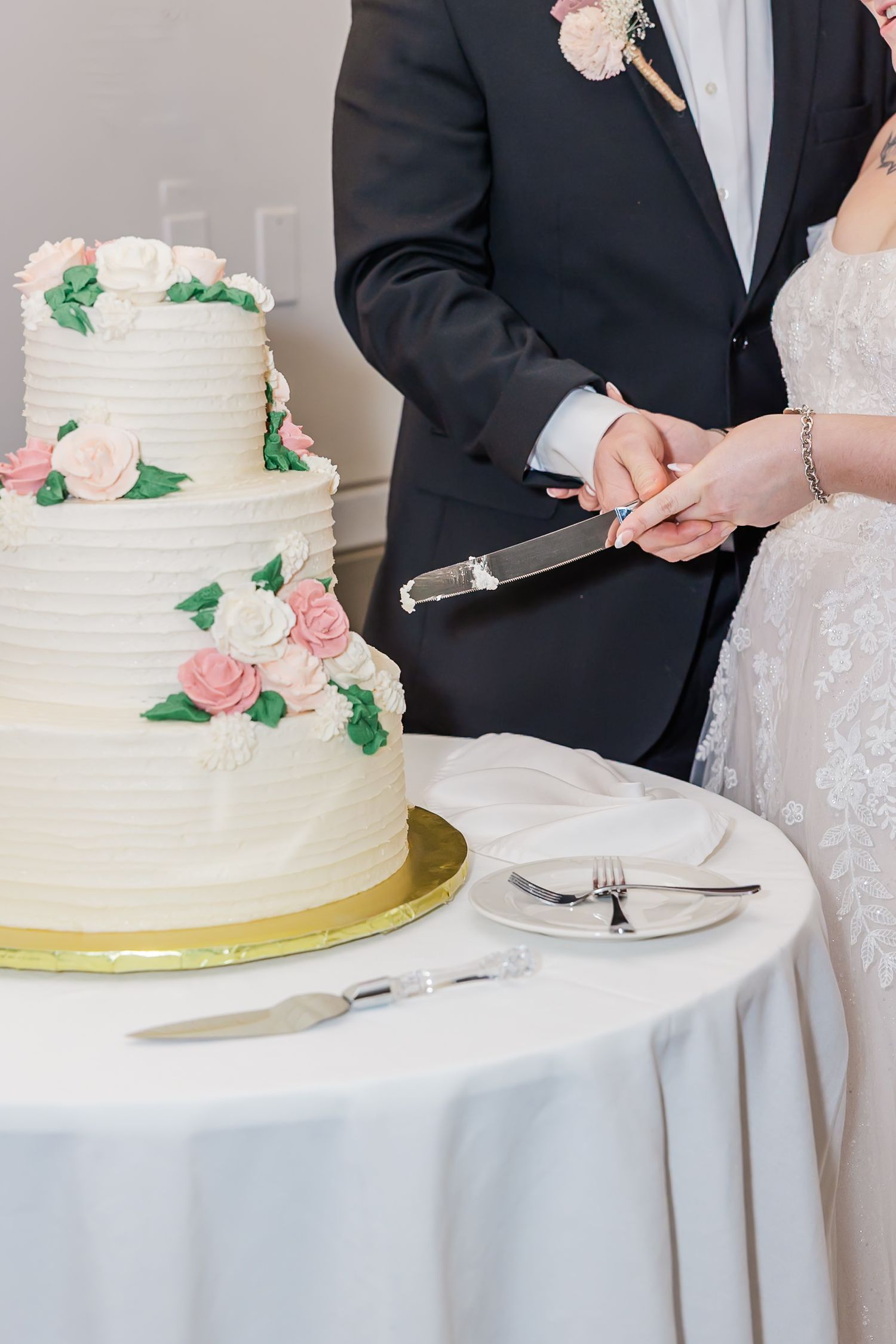 A wedding cake decorated with pink roses being cut on a white tablecloth at a reception.