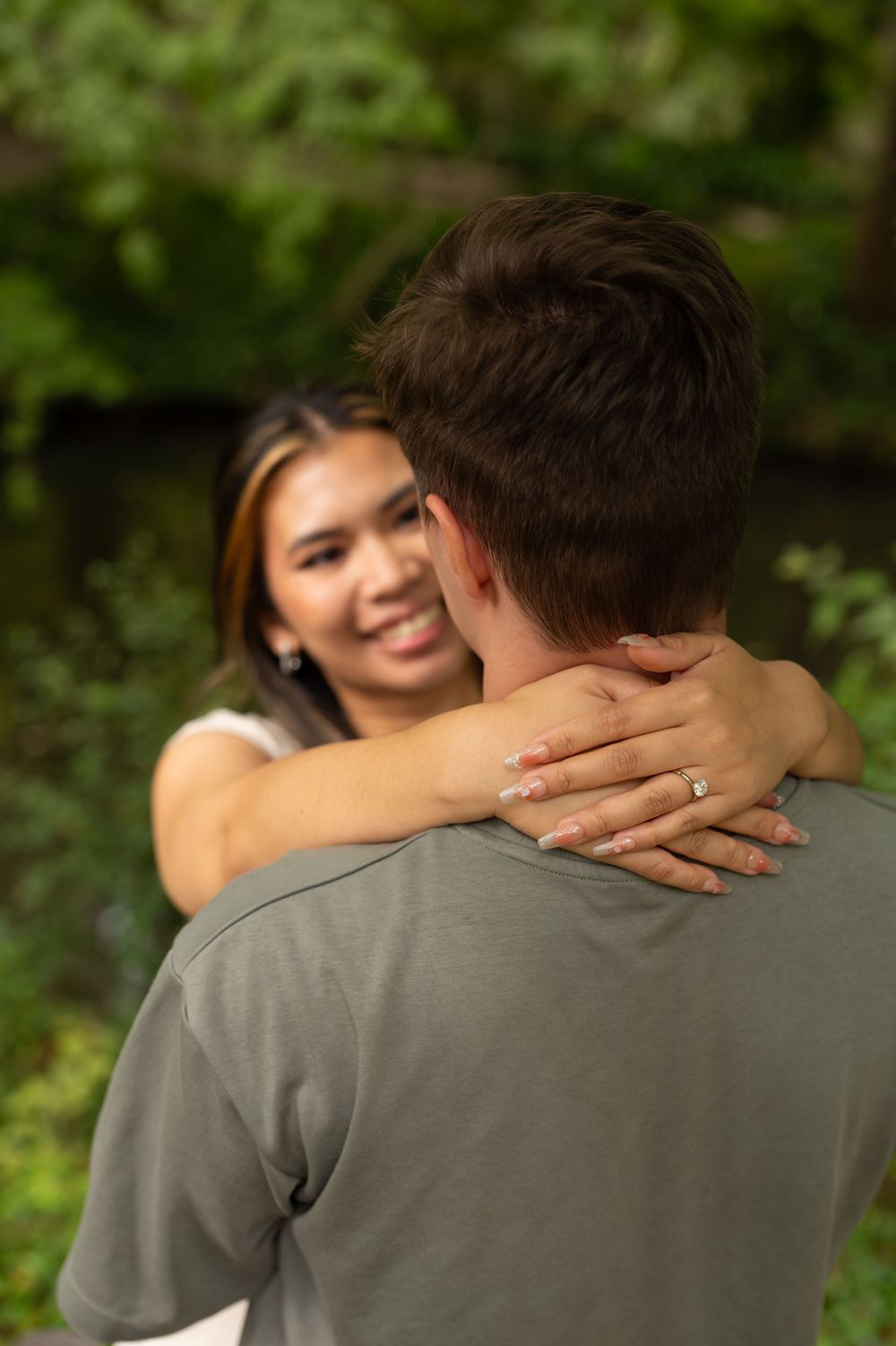 Woman embracing man from behind, displaying engagement ring, in a lush green outdoor setting.