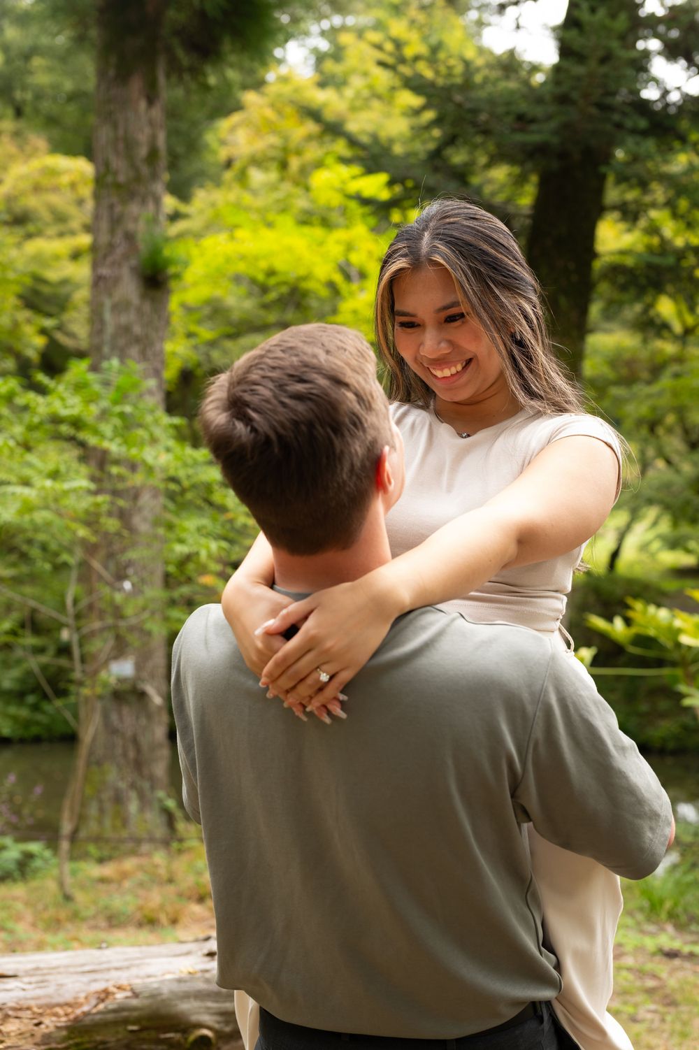 Woman smiling joyfully while being lifted by her partner in a lush green Japanese garden.