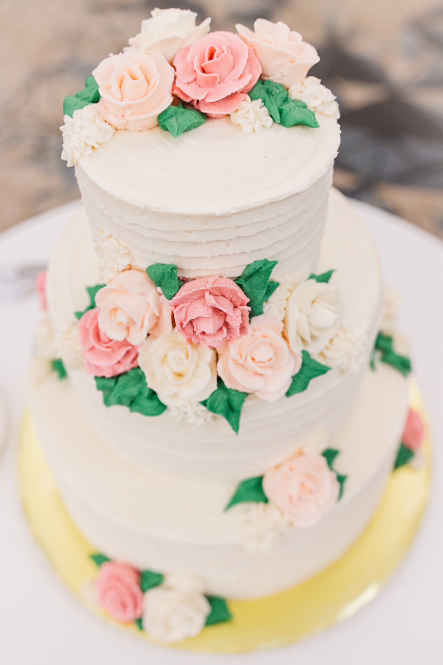 A white wedding cake decorated with delicate pink roses and green leaves sits on a yellow plate.
