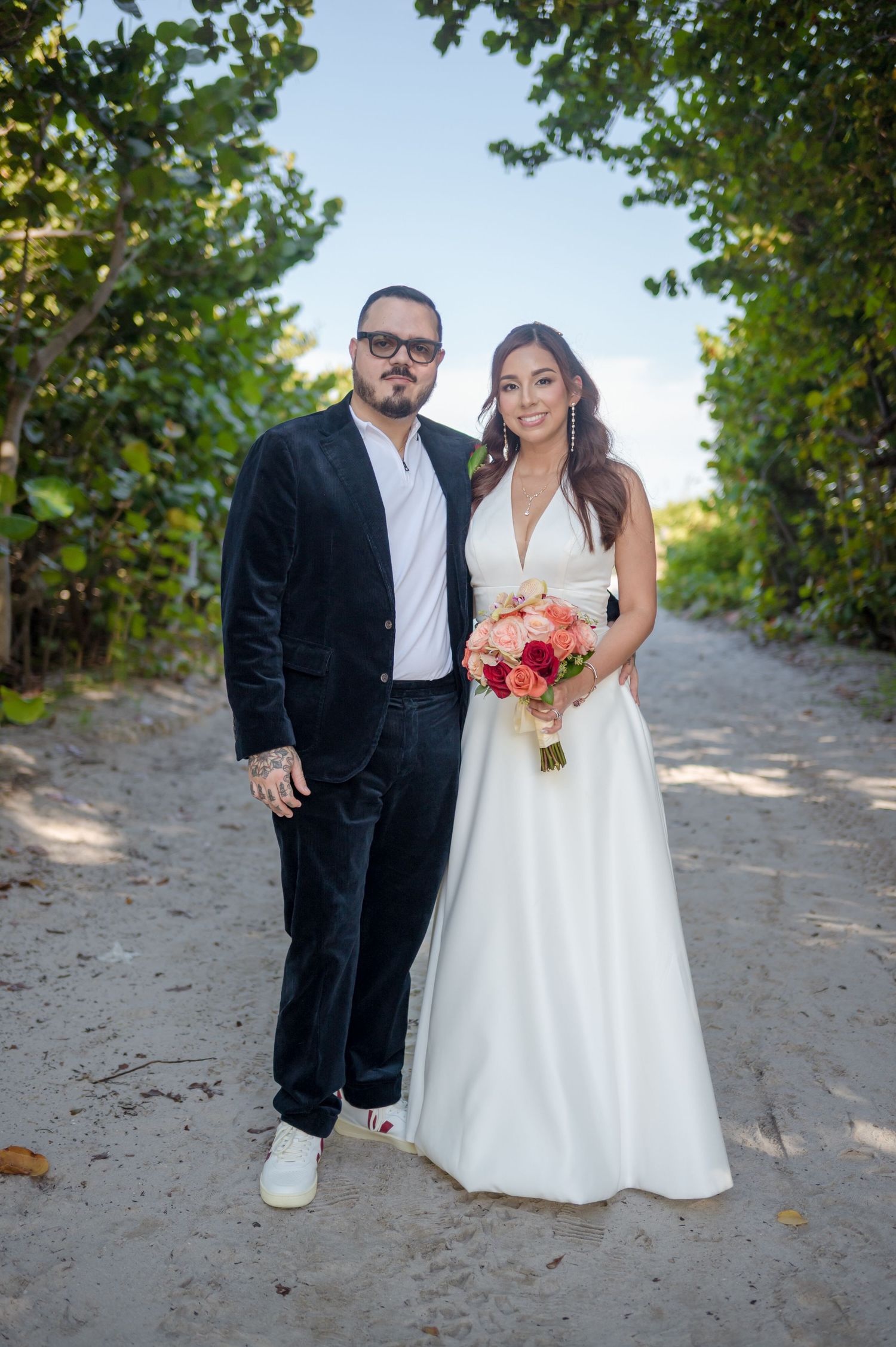 A couple poses together on a sandy beach path surrounded by green foliage during their wedding photoshoot.