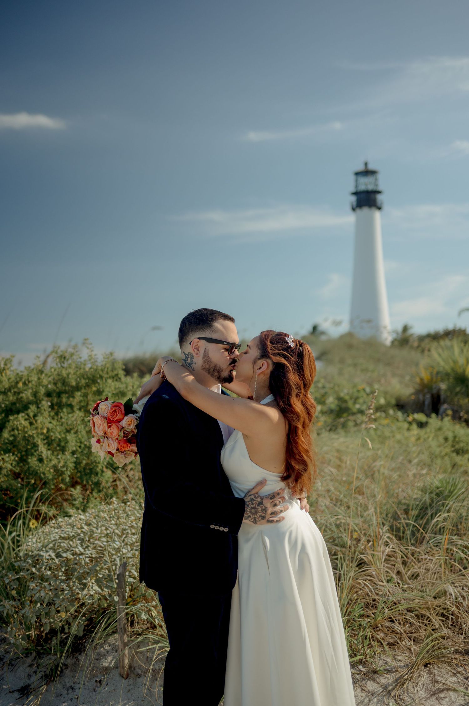 A lighthouse stands tall against a cloudy sky while a couple shares a romantic moment in the grassy dunes.