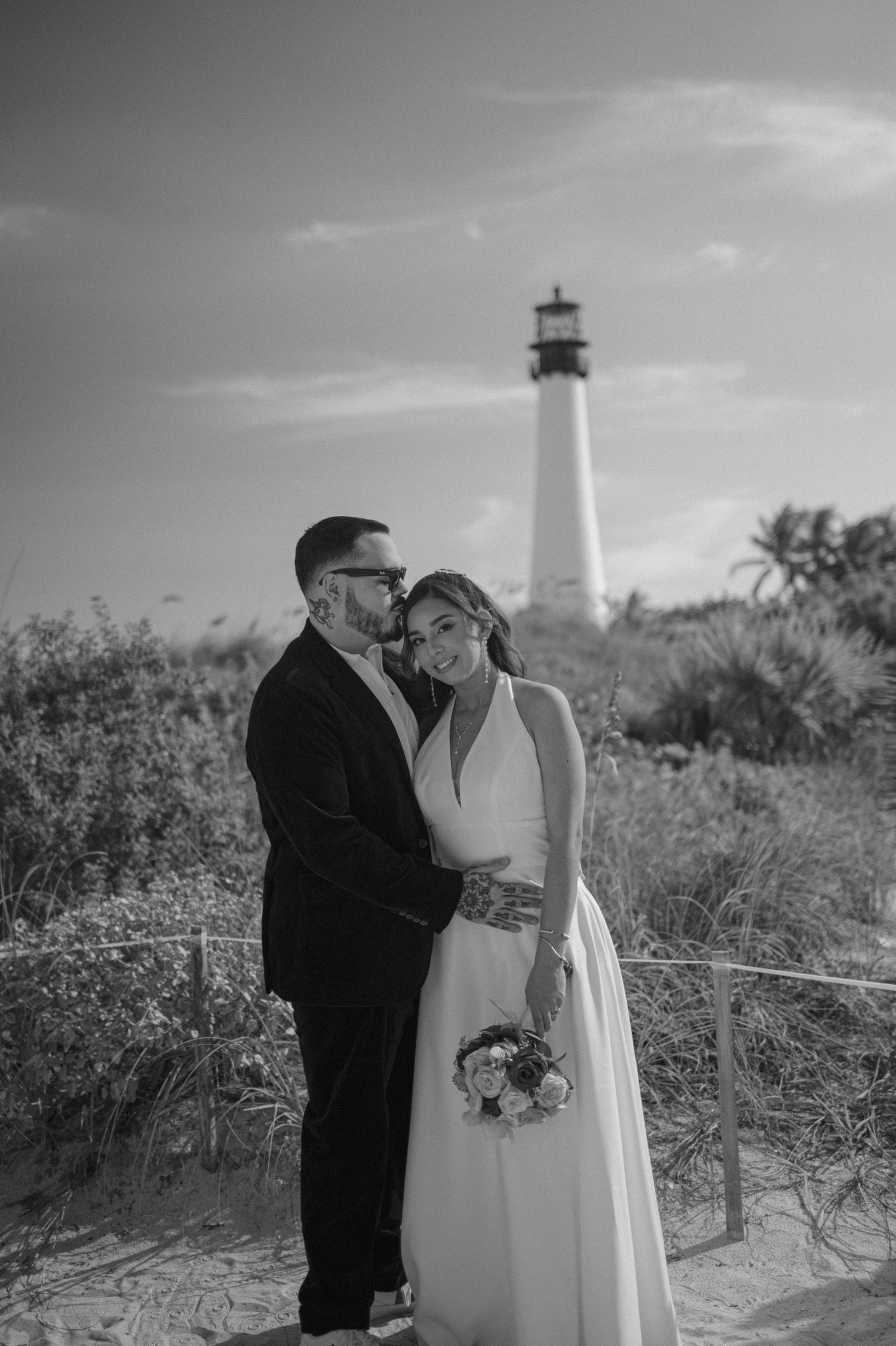 Black and white wedding photos show couple embracing near a lighthouse on a coastal beach with tall grass.