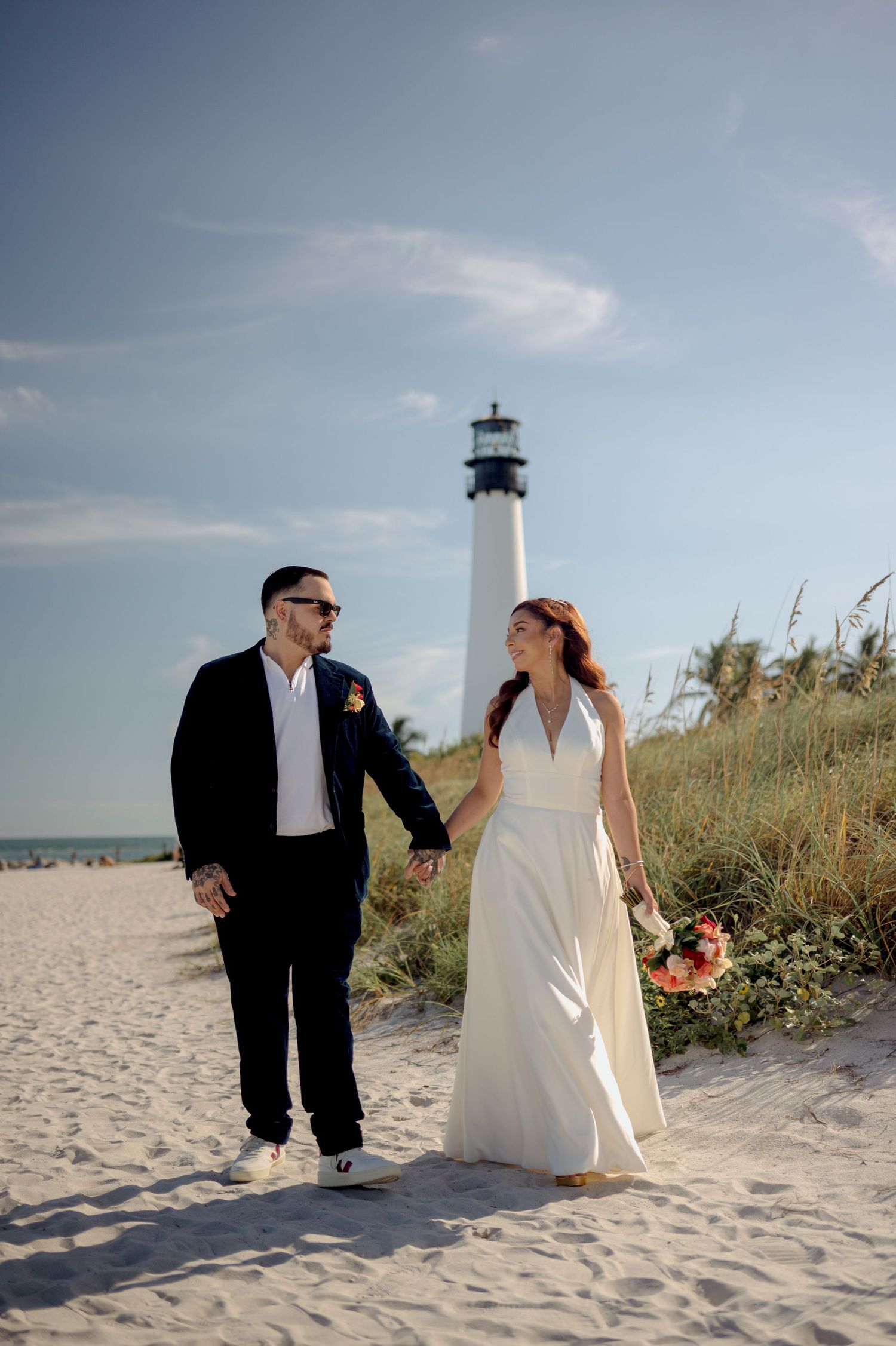 A couple in wedding attire walking along a sandy beach with sea oats and ocean views during a beach wedding photoshoot.