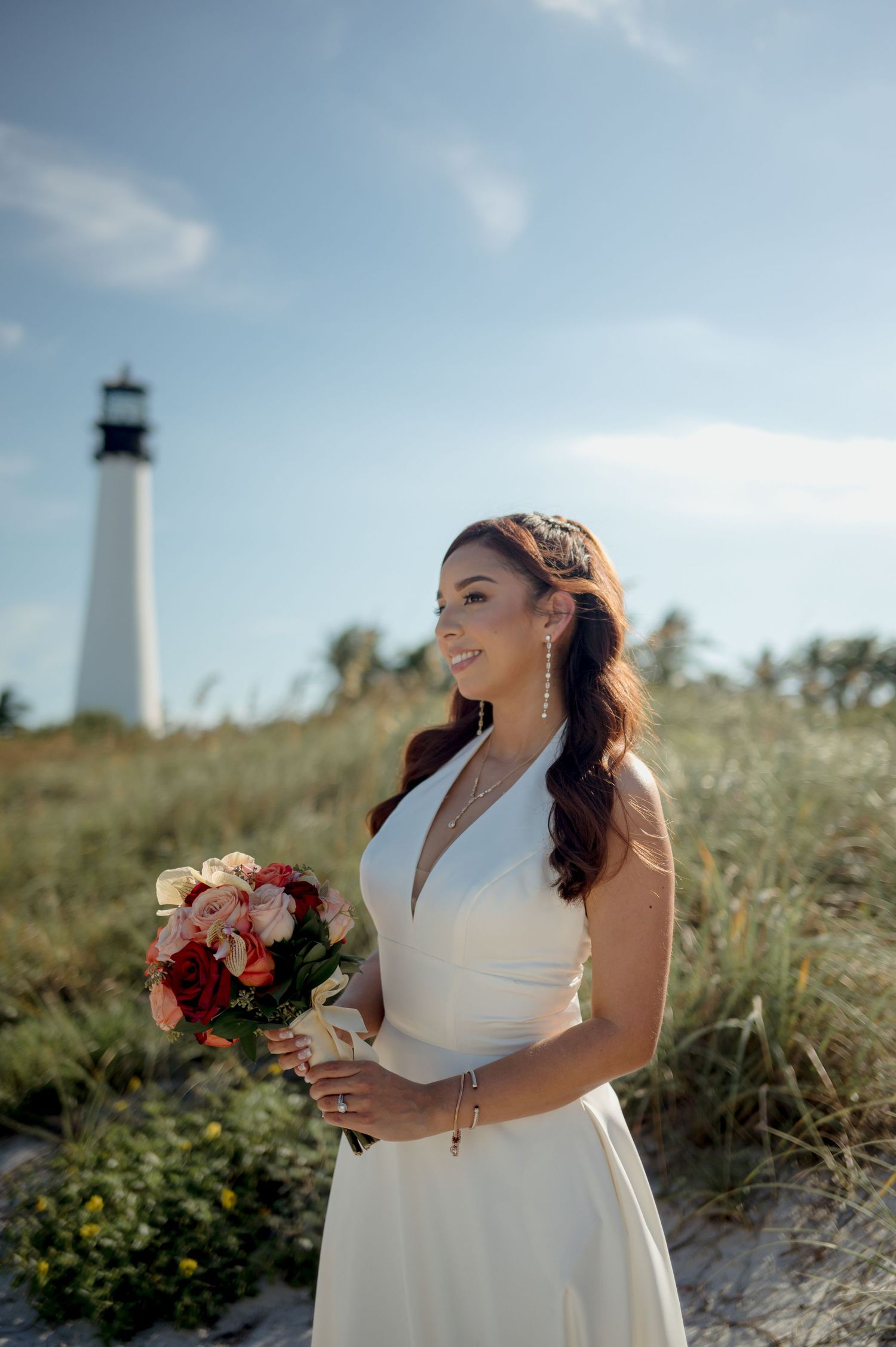 A lighthouse stands tall in the background as someone in a white wedding dress holds a bouquet of red and pink flowers.