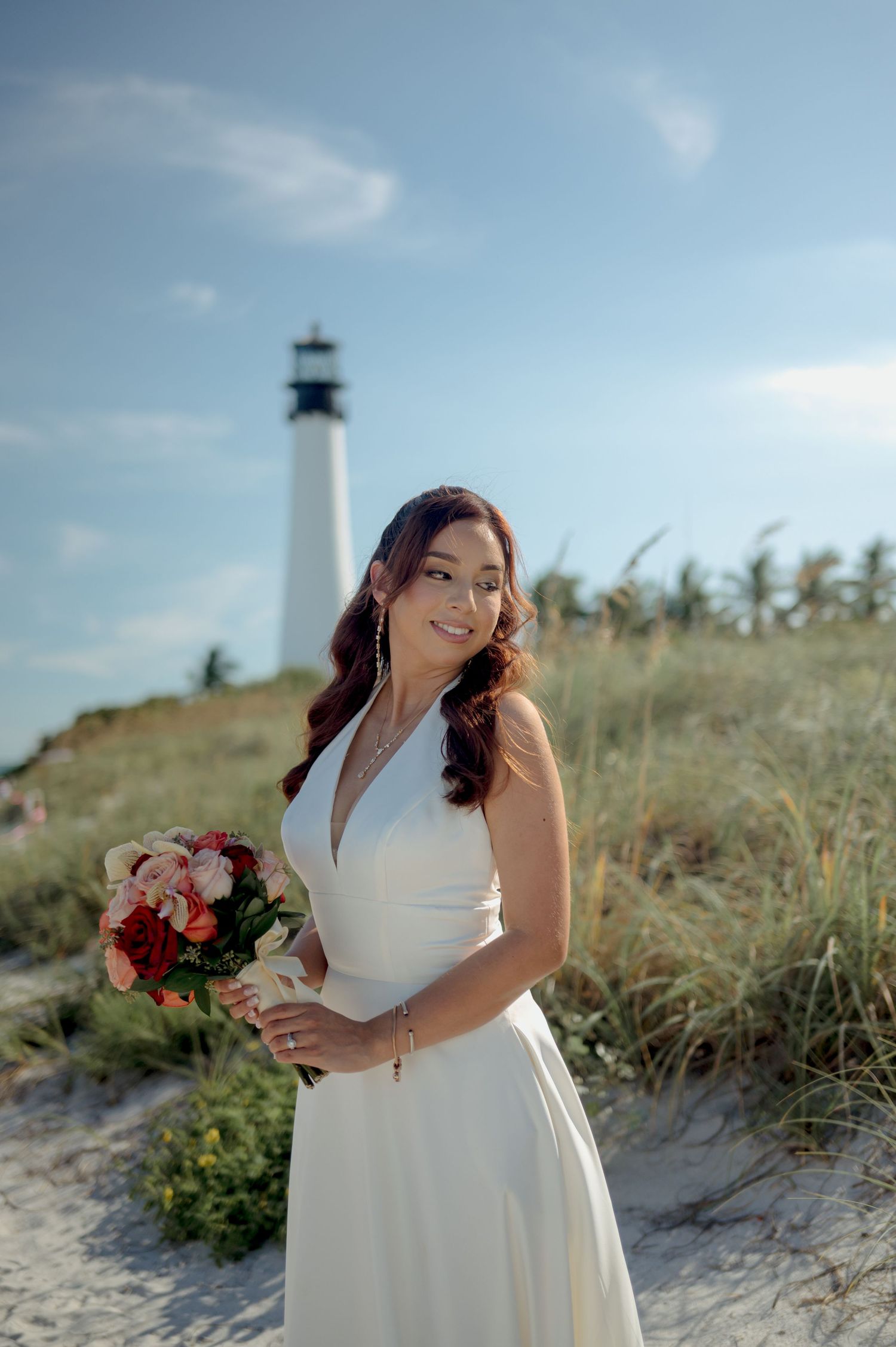 A lighthouse stands tall in the background as someone in a white wedding dress holds a colorful bouquet on a sunny beach.