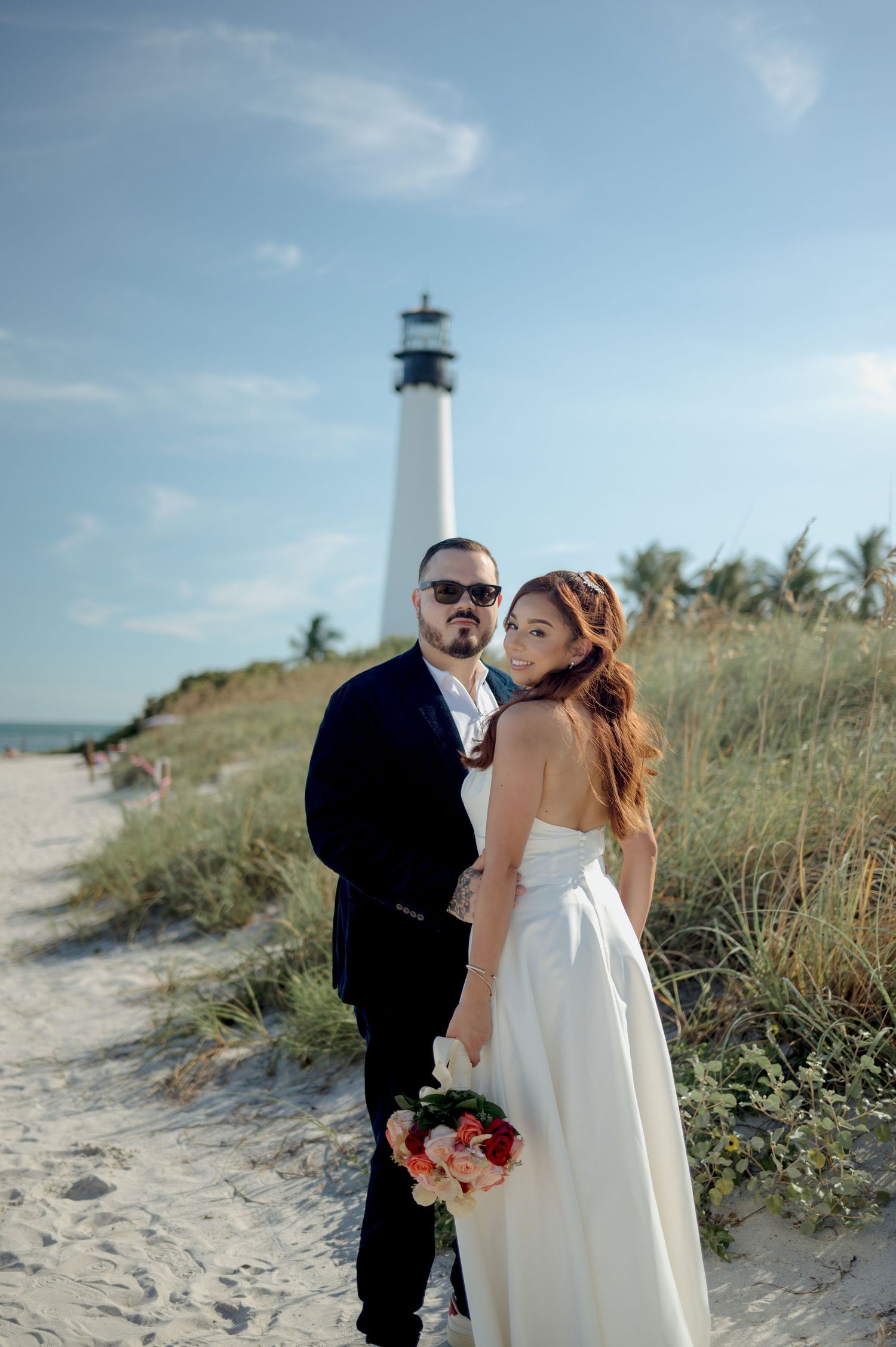 A couple in wedding attire poses together on a sandy beach path with a lighthouse and palm trees in the background.