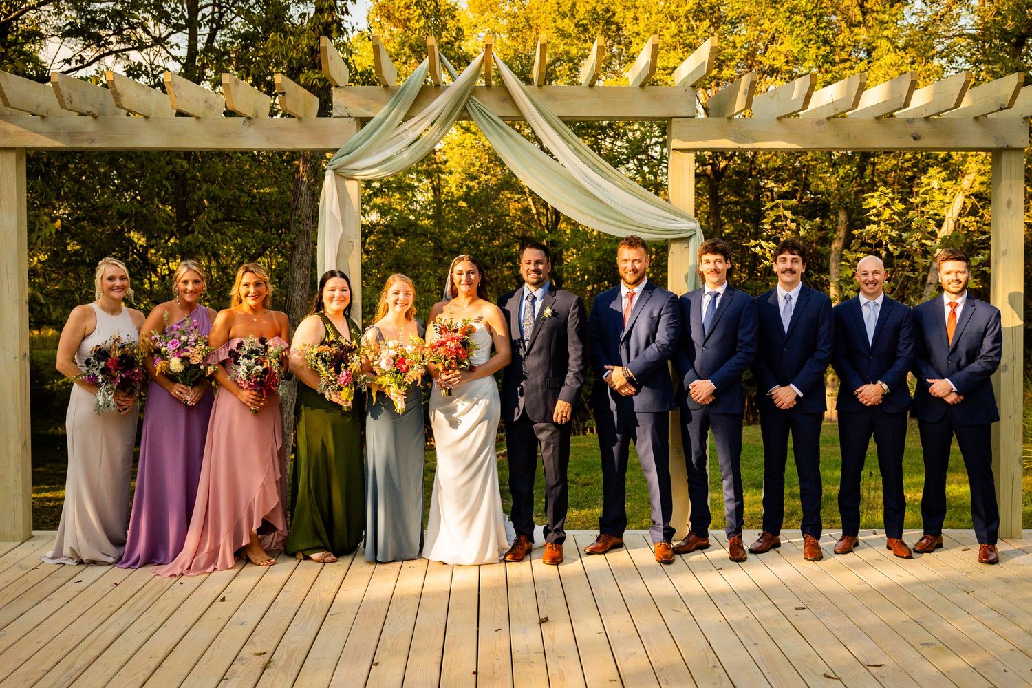 Wedding party stands in line under wooden pergola with white draping and autumn foliage in background.