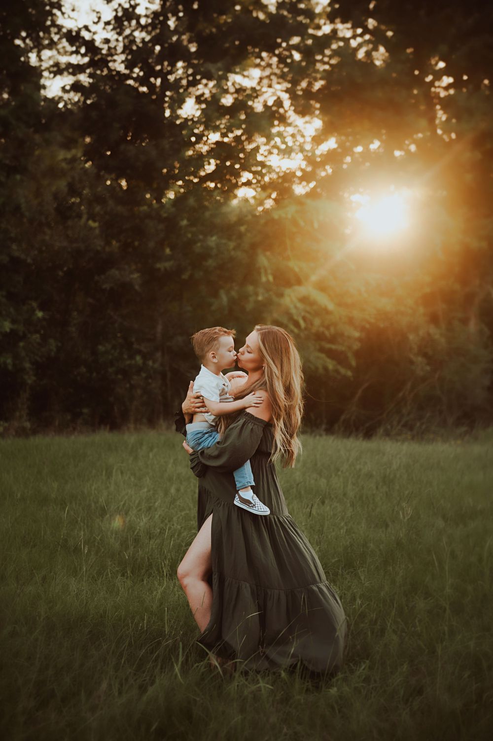 A mother in a green dress kisses her toddler son at golden hour in a lush meadow.