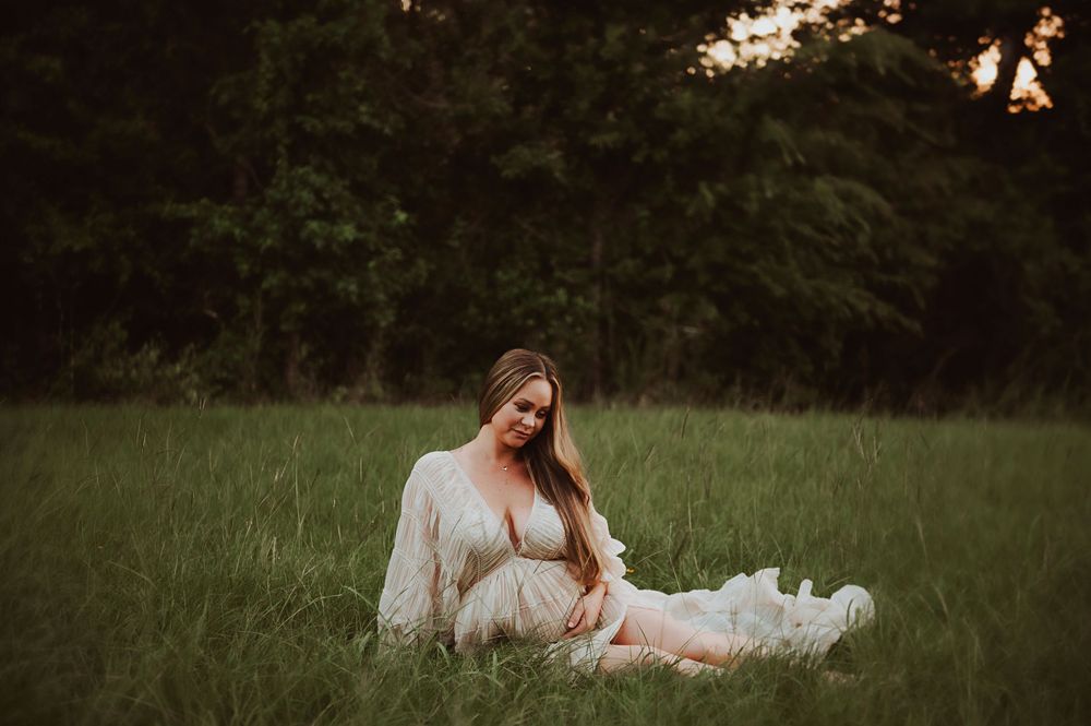 Pregnant woman in white lace dress sitting in green field at sunset, cradling baby bump.