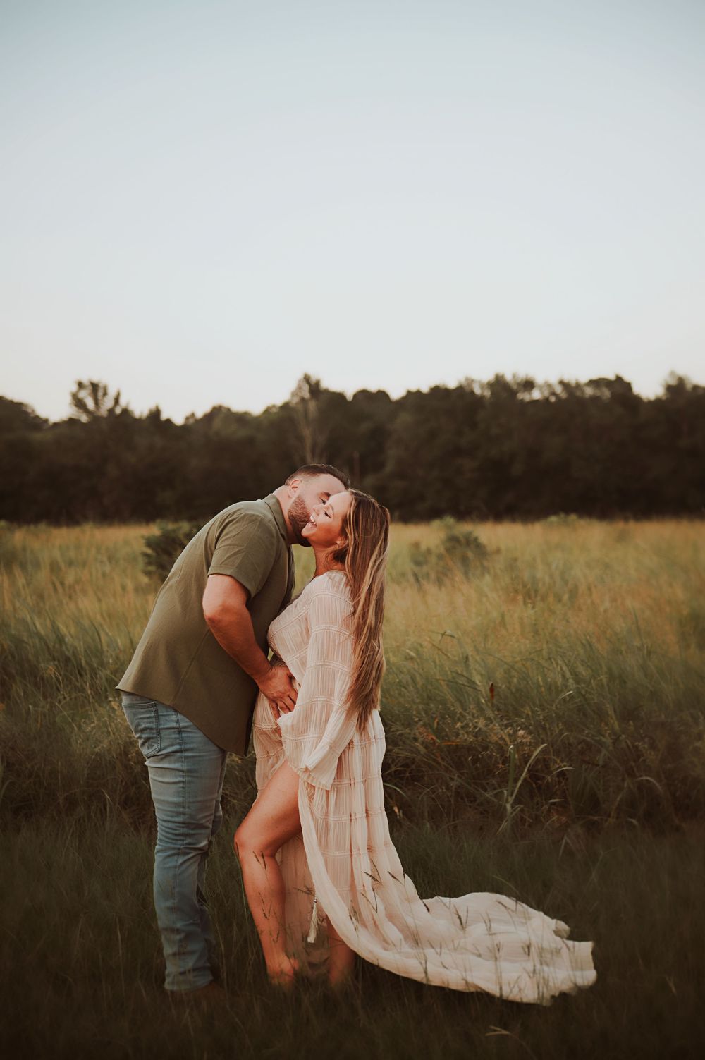 A couple shares a romantic kiss in a sunlit meadow, the woman wearing a flowing white gown as golden hour light glows behind them.