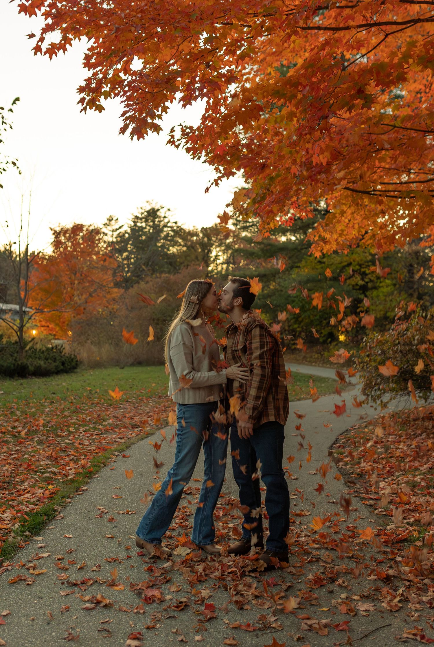 A couple shares a romantic moment on a path covered in vibrant autumn leaves under orange maple trees.