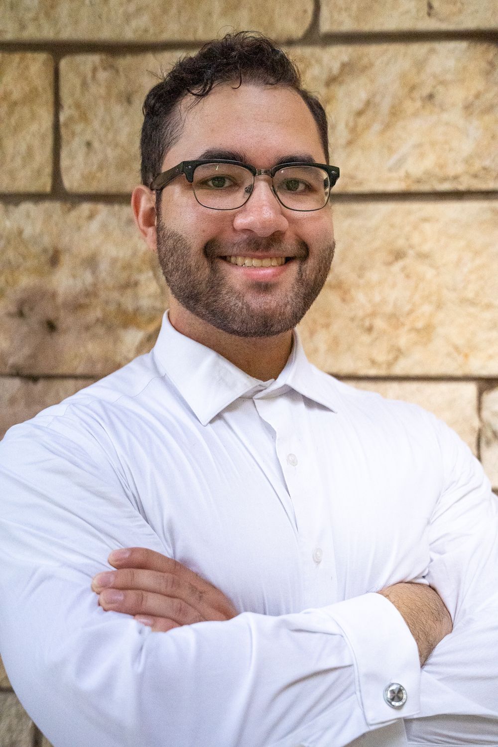 A professional in a white dress shirt and glasses poses confidently against a stone wall.