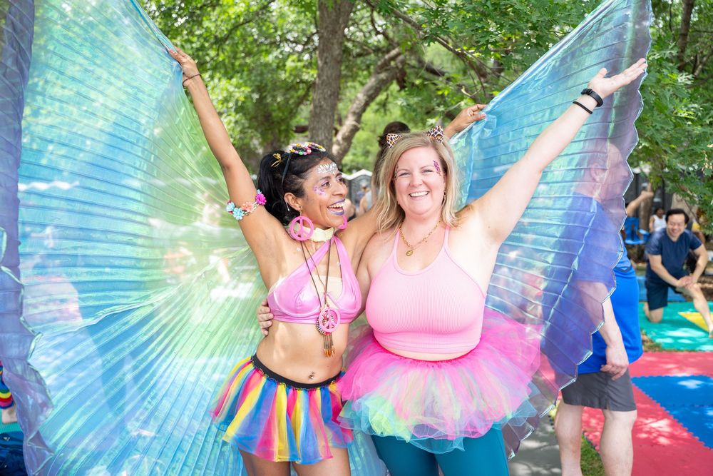 Two festival-goers wearing colorful fairy wings and tutus celebrate joyfully at an outdoor summer event.