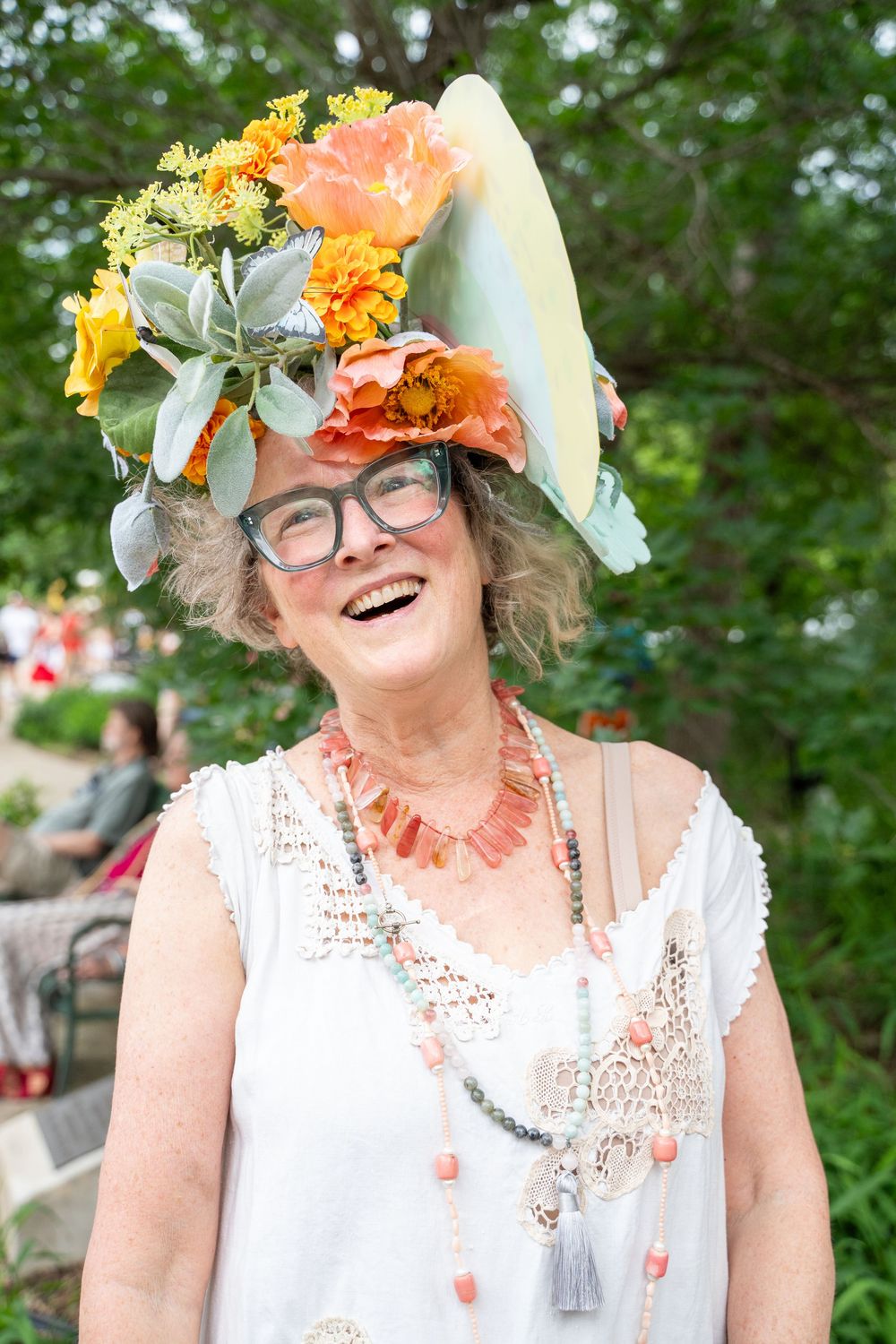 A joyful figure wearing glasses and a floral crown smiles brightly in a white dress at an outdoor summer event.