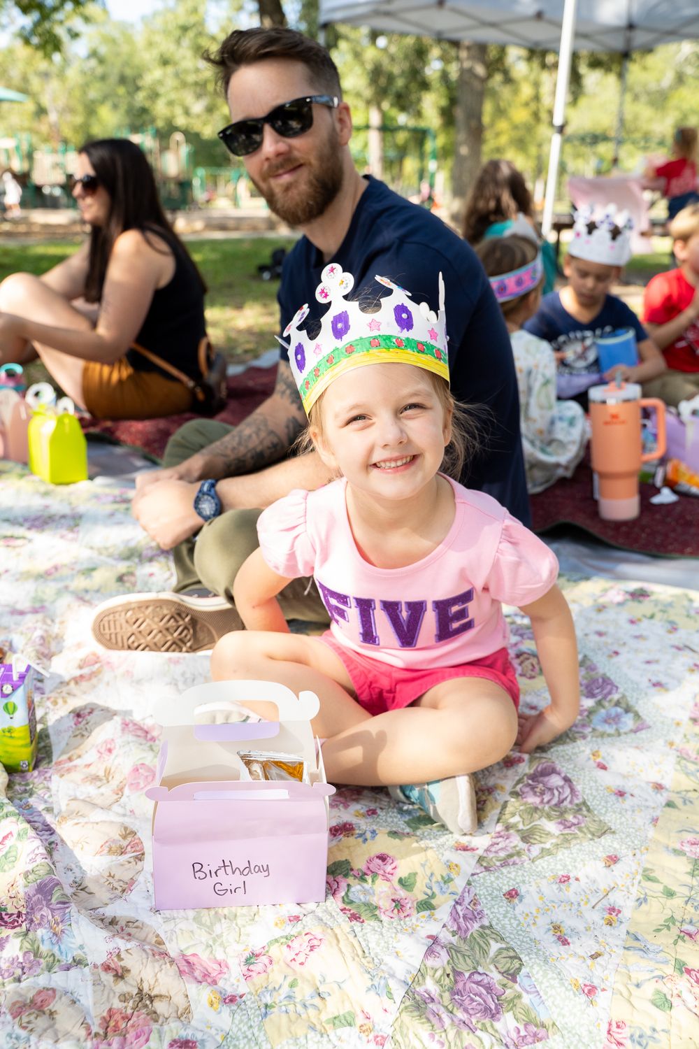 A smiling girl wearing a crown and FIVE shirt sits with her Birthday Girl box at an outdoor birthday party.