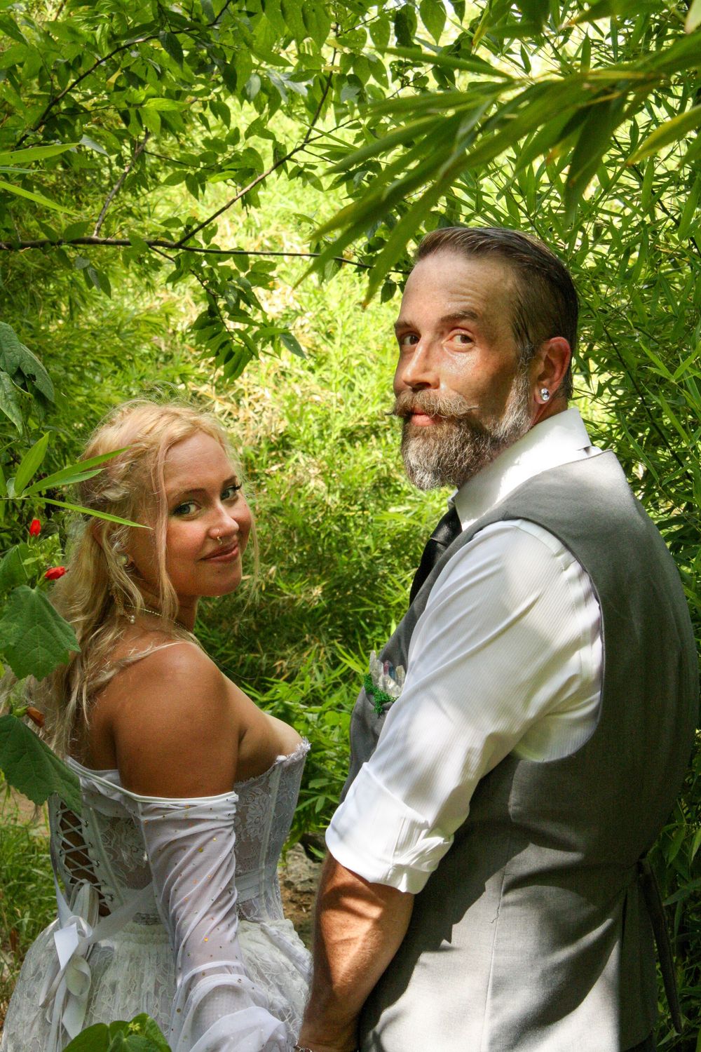 A couple in wedding attire poses together under lush green foliage in a garden setting.