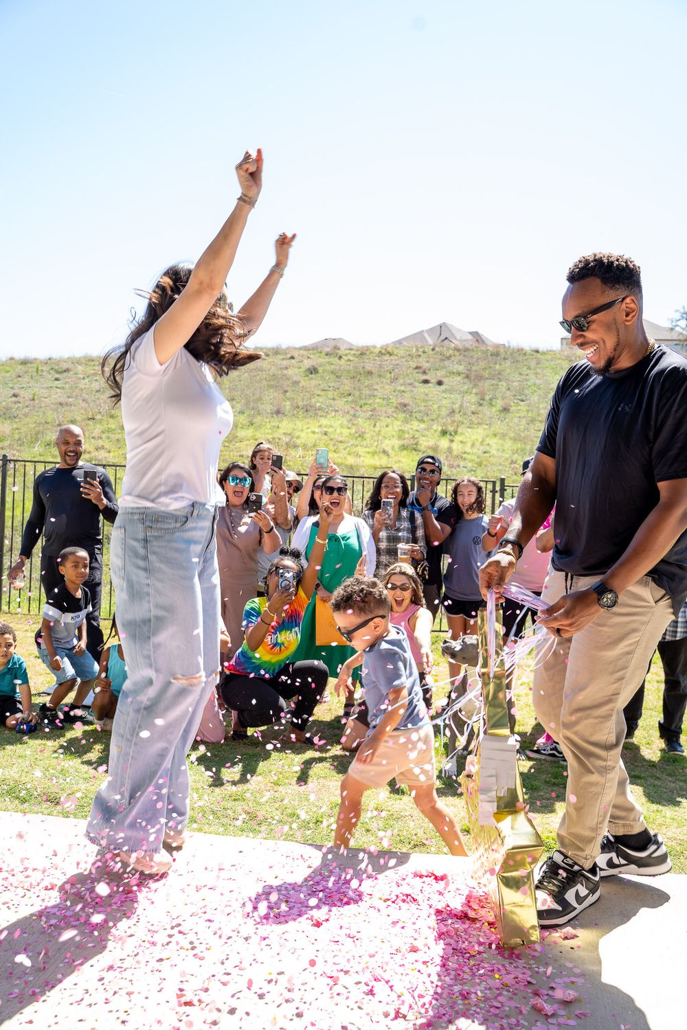 Family celebrating a gender reveal party outdoors with pink confetti, cheering crowd, and sunny hillside backdrop.