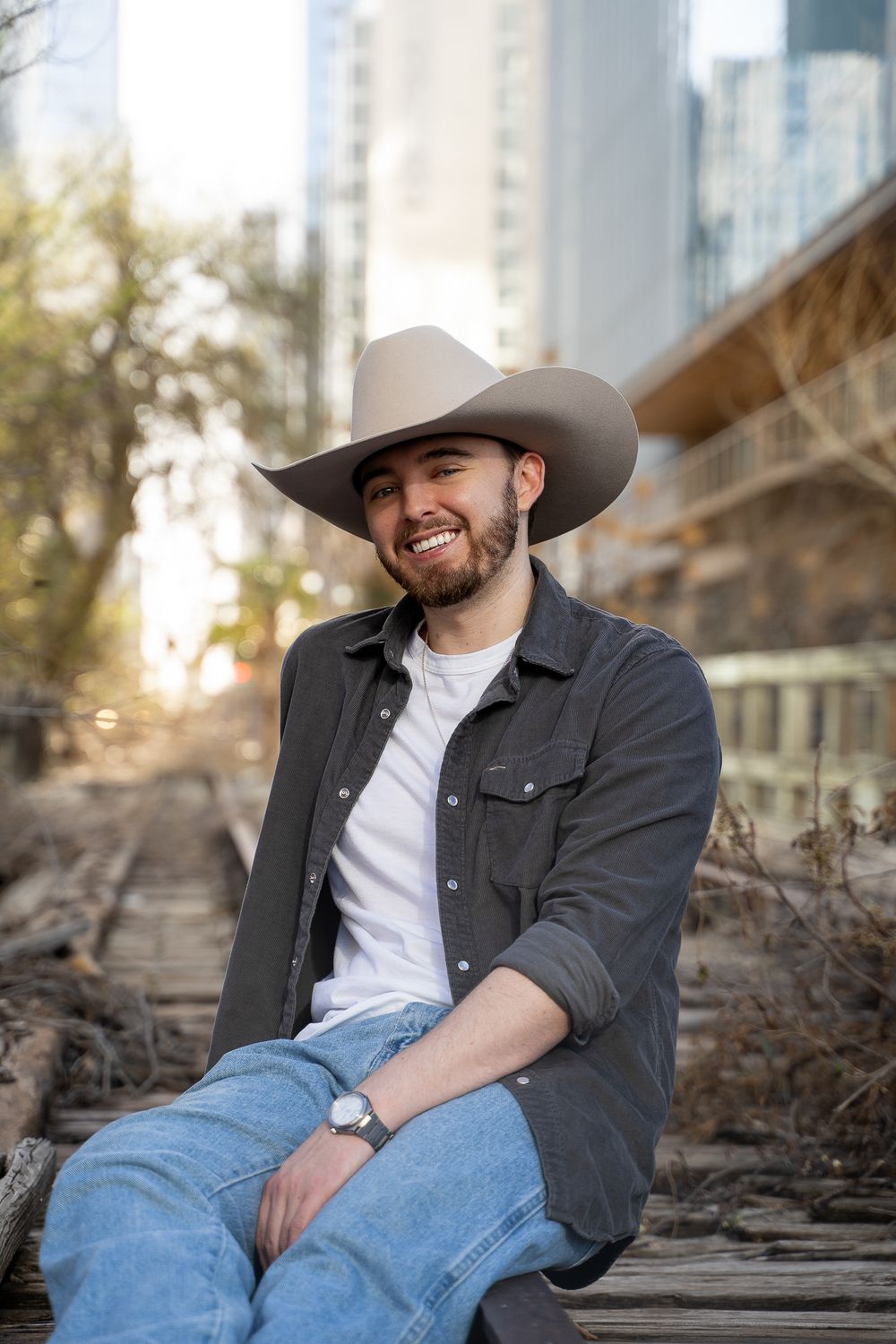 Smiling bearded man in cowboy hat, dark shirt, and jeans sitting on railroad tracks with city buildings in background.