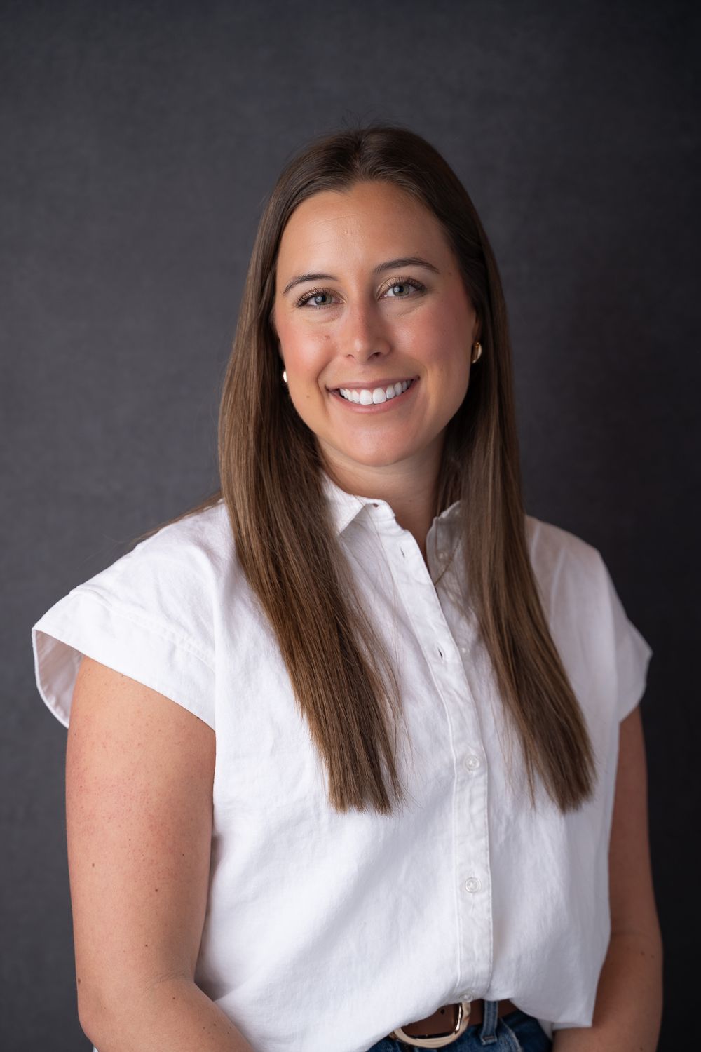 Smiling woman with long brown hair wearing a white sleeveless button-up shirt against a dark gray background.