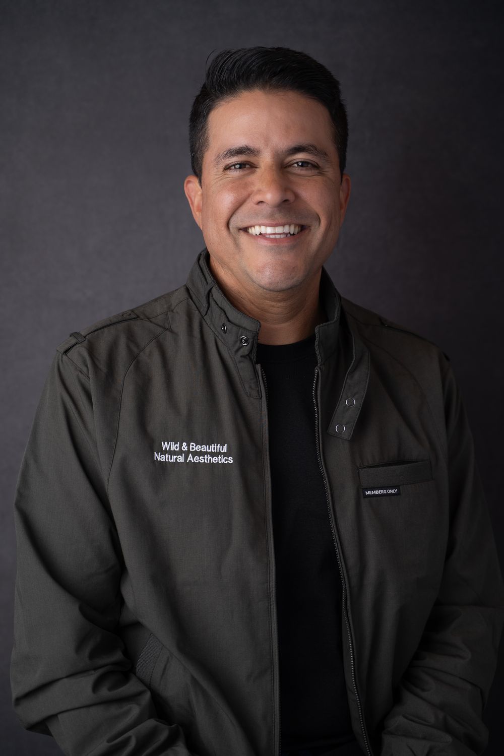 Smiling man wearing a dark olive jacket with Wild & Beautiful Natural Aesthetics logo against a gray background.