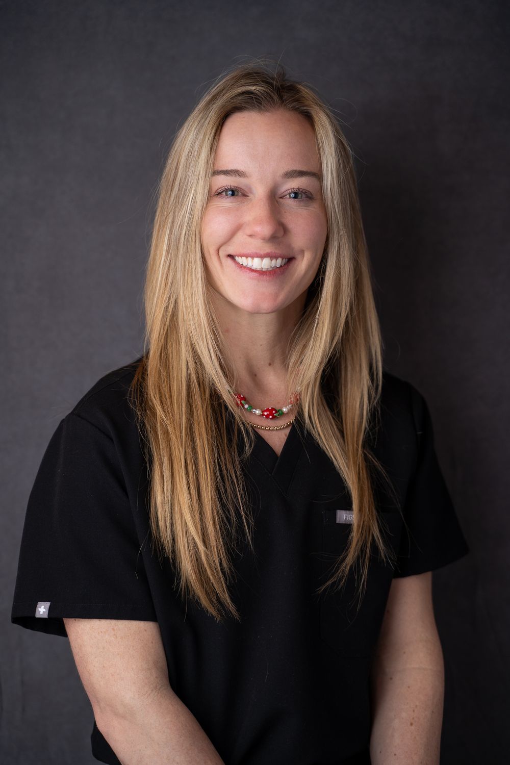 Smiling female healthcare professional with long blonde hair wearing black scrubs and a red beaded necklace.