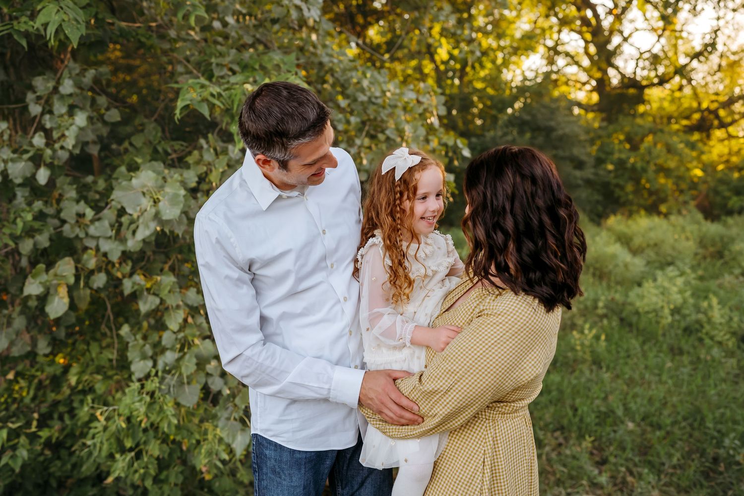 A couple embraces outdoors during golden hour in a natural setting with trees and sunlight filtering through the foliage.