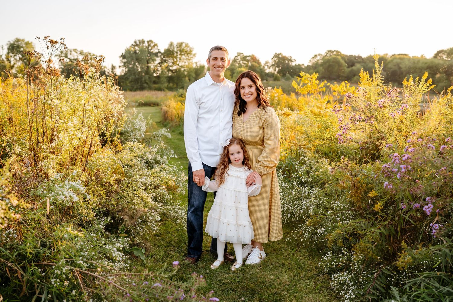 A family stands together in a golden meadow at sunset surrounded by wildflowers and tall grass.