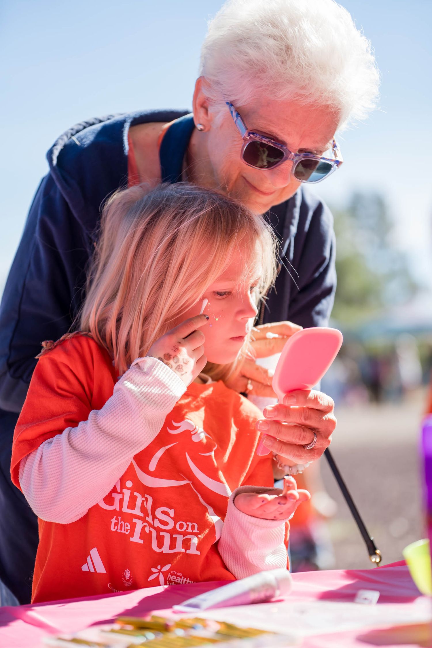 A child in a red shirt looks at a mobile device while an adult with white hair and sunglasses leans in to watch.