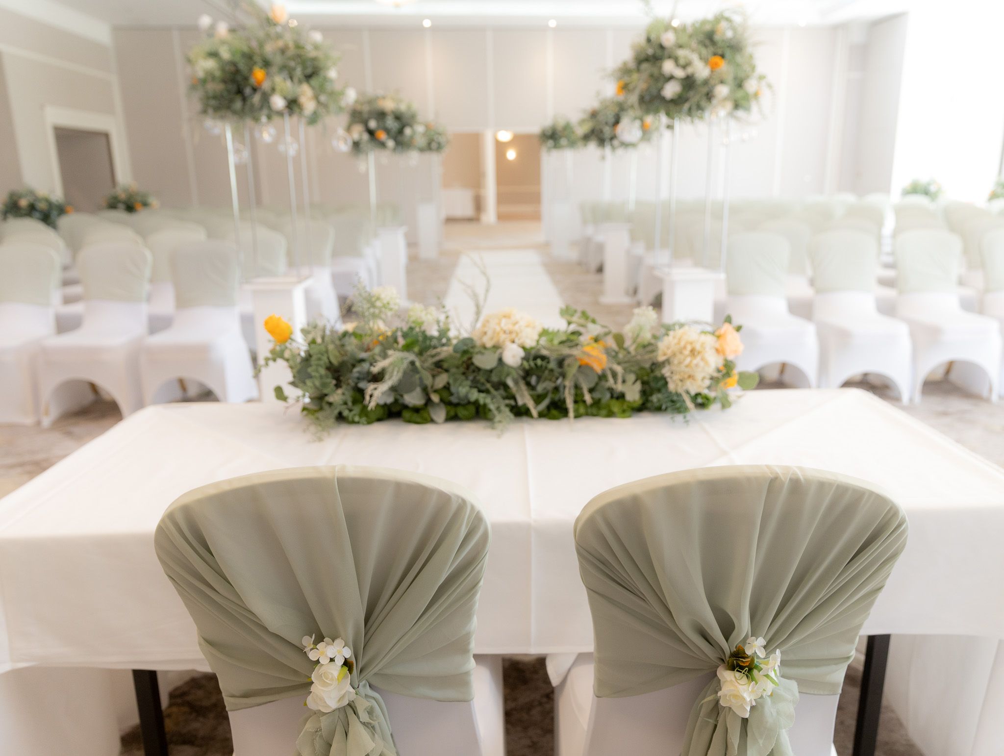 Elegant white wedding reception tables decorated with floral centerpieces and draped chair covers in a bright banquet hall.