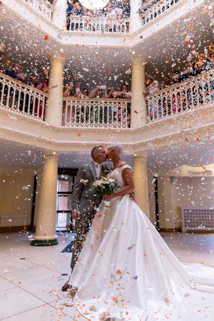 Bride and groom twirl under falling confetti in an elegant white columned ballroom during their first dance.