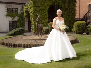 A bride in a flowing white wedding dress poses on a grassy lawn beside a brick building.