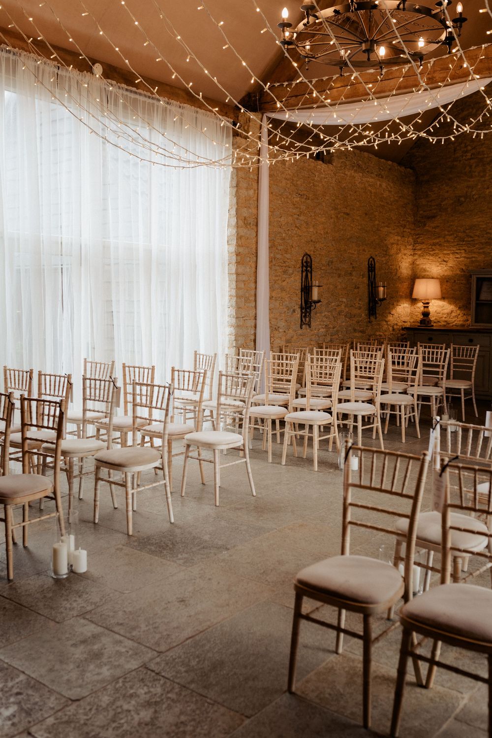 Rustic wedding venue interior with rows of white Chiavari chairs and twinkling string lights on exposed brick walls.