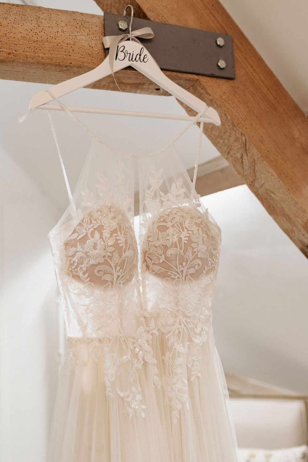 Delicate blush wedding dress with lace details hanging on white hanger against rustic wooden beams.